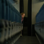 A still from the Apple TV+ series Pluribus, showing actress Rhea Seehorn as the character Carol looking anxious while hiding behind a row of blue airplane seats in a dimly lit cabin, illustrating the theme of isolation.
