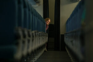 A still from the Apple TV+ series Pluribus, showing actress Rhea Seehorn as the character Carol looking anxious while hiding behind a row of blue airplane seats in a dimly lit cabin, illustrating the theme of isolation.